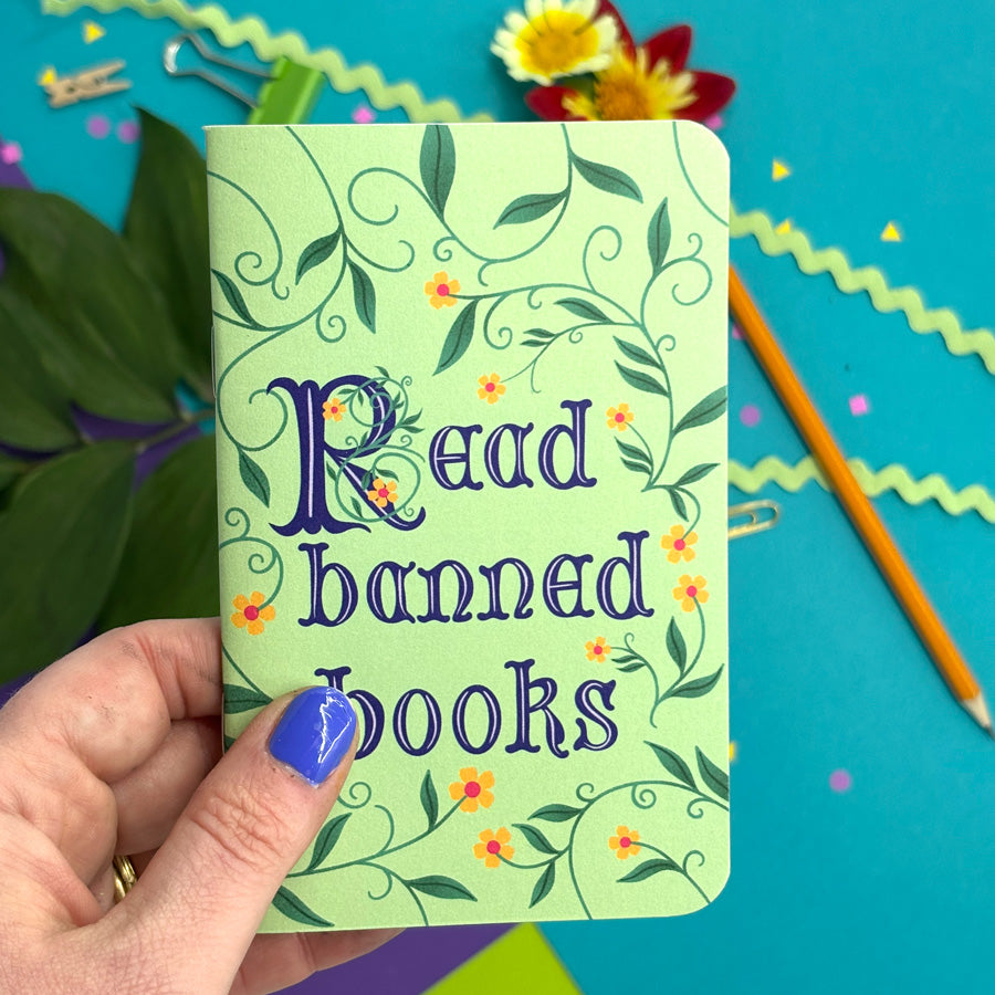 hand with blue nail polish holding small green pocket notebook with medieval lettering that says "read banned books" with floral illustrations
