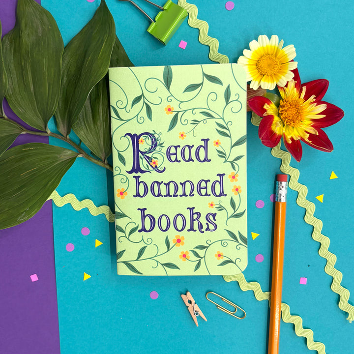 A small notebook with a pale green cover decorated with green vines and small orange flowers, featuring the phrase 'Read banned books' in purple lettering. The book is styled in a flat-lay arrangement on a bright blue background with decorative paper, a pencil, binder clips, leaves, and flowers.