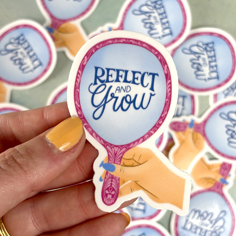 hand holding A sticker with a mirror design and the text 'Reflect and Grow' printed on it in a light-colored font, predominantly pink and blue in color.