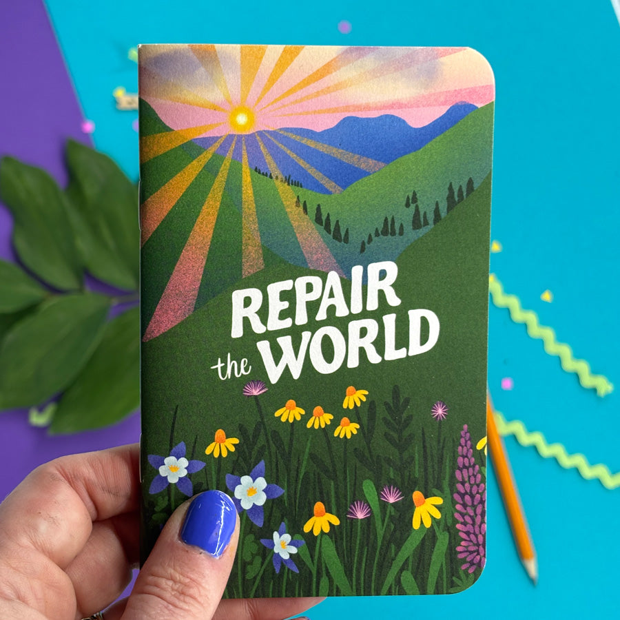 A hand with blue nail polish holds a small notebook featuring a colorful landscape illustration with the text 'REPAIR the WORLD' in white. The background is a bright turquoise surface with decorative paper elements and a pencil.