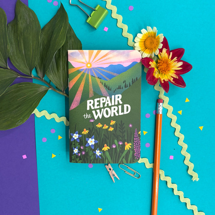 A  notebook with a colorful landscape cover reading 'REPAIR the WORLD' is arranged in a styled flat lay on a bright turquoise background with paper clips, a pencil, flowers, leaves, and confetti.