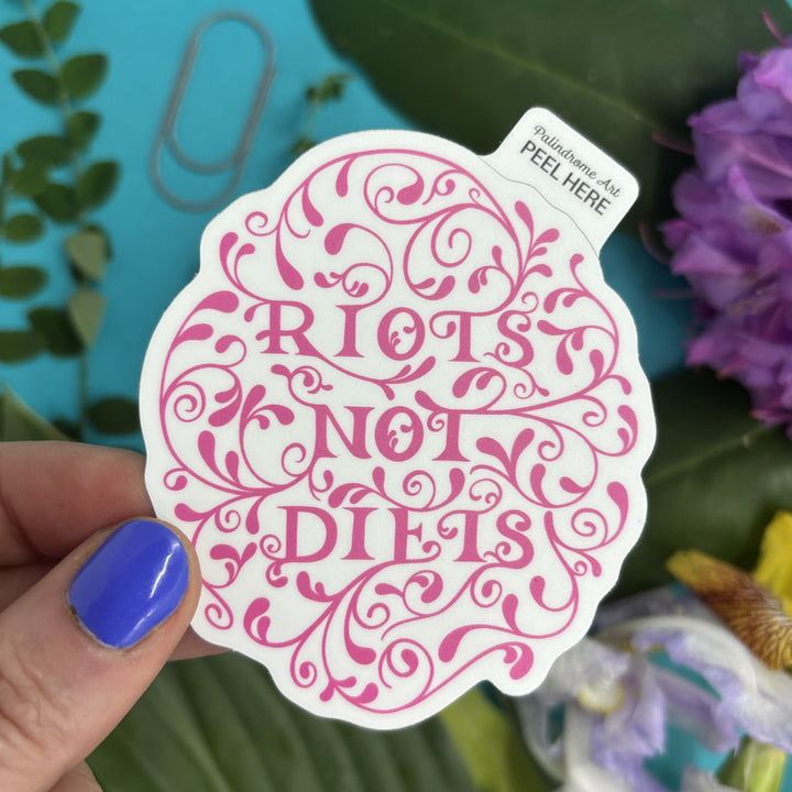 Sticker with 'Riots Not Diets' text held by a hand with purple nail polish, against a blurred natural background.