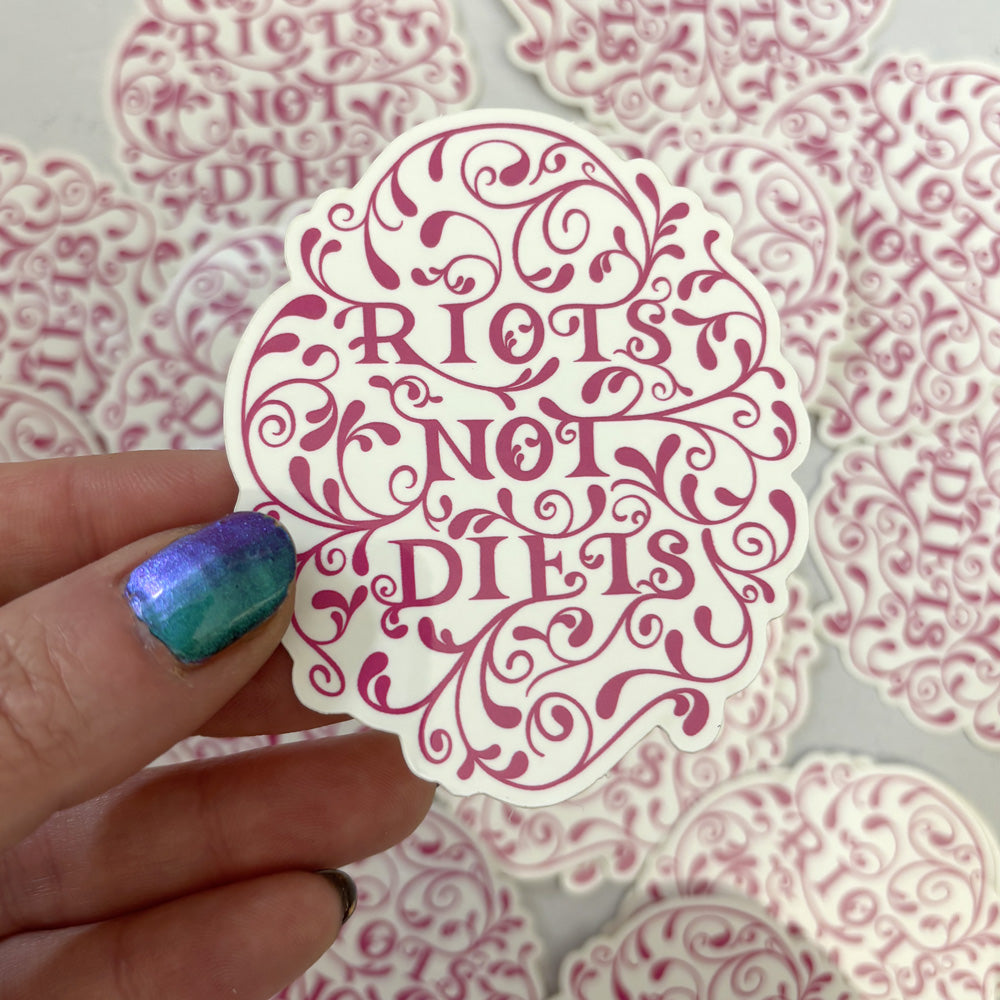 Sticker with 'Riots Not Diets' text held by a hand against a patterned background