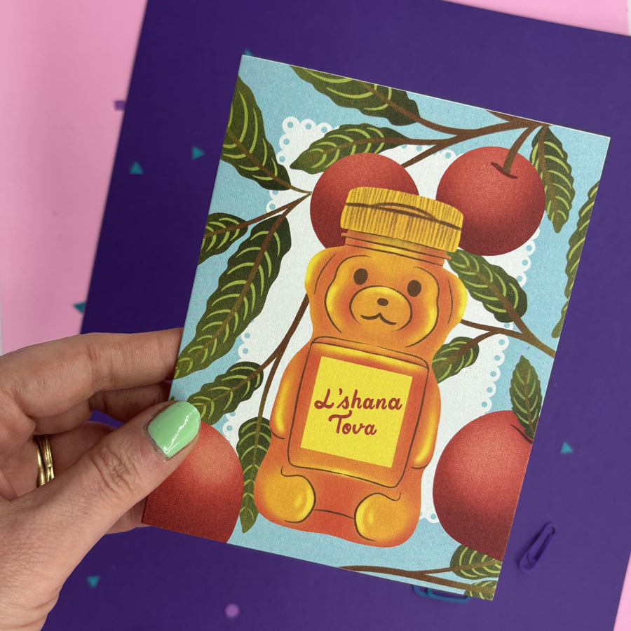 A hand with mint green nail polish holds a Rosh Hashana greeting card featuring a honey bear labeled 'Lashana Tova' surrounded by red apples on a light blue background. 