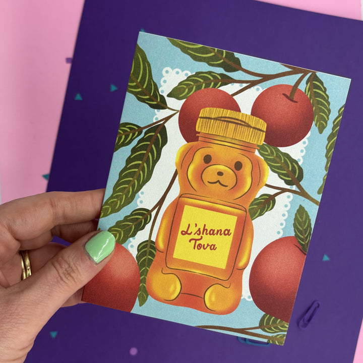 A hand with mint green nail polish holds a Rosh Hashana greeting card featuring a honey bear labeled 'Lashana Tova' surrounded by red apples on a light blue background. 
