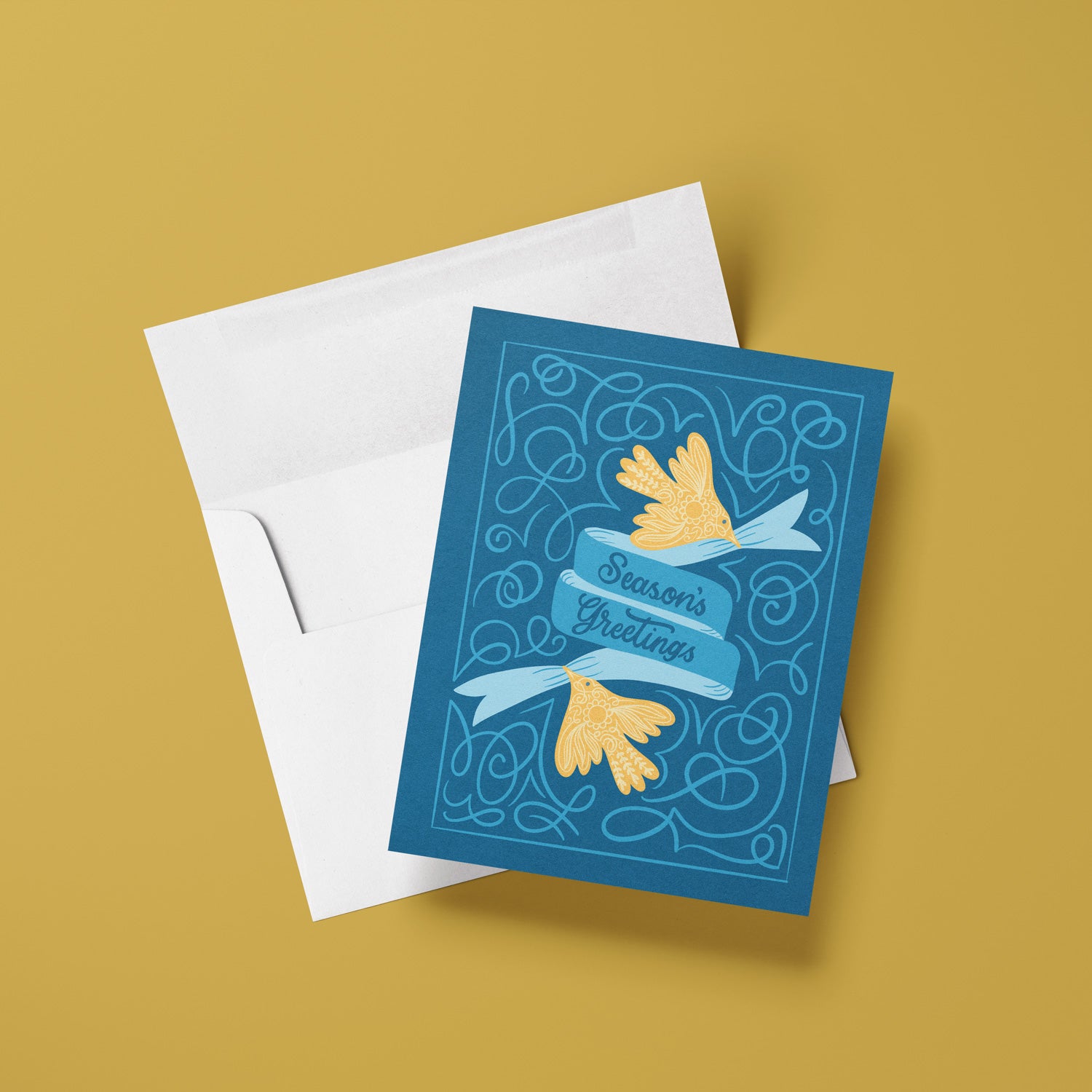 Greeting card with 'Season's Greetings' text and gold birds on a blue background, with a white envelope on a mustard yellow background.