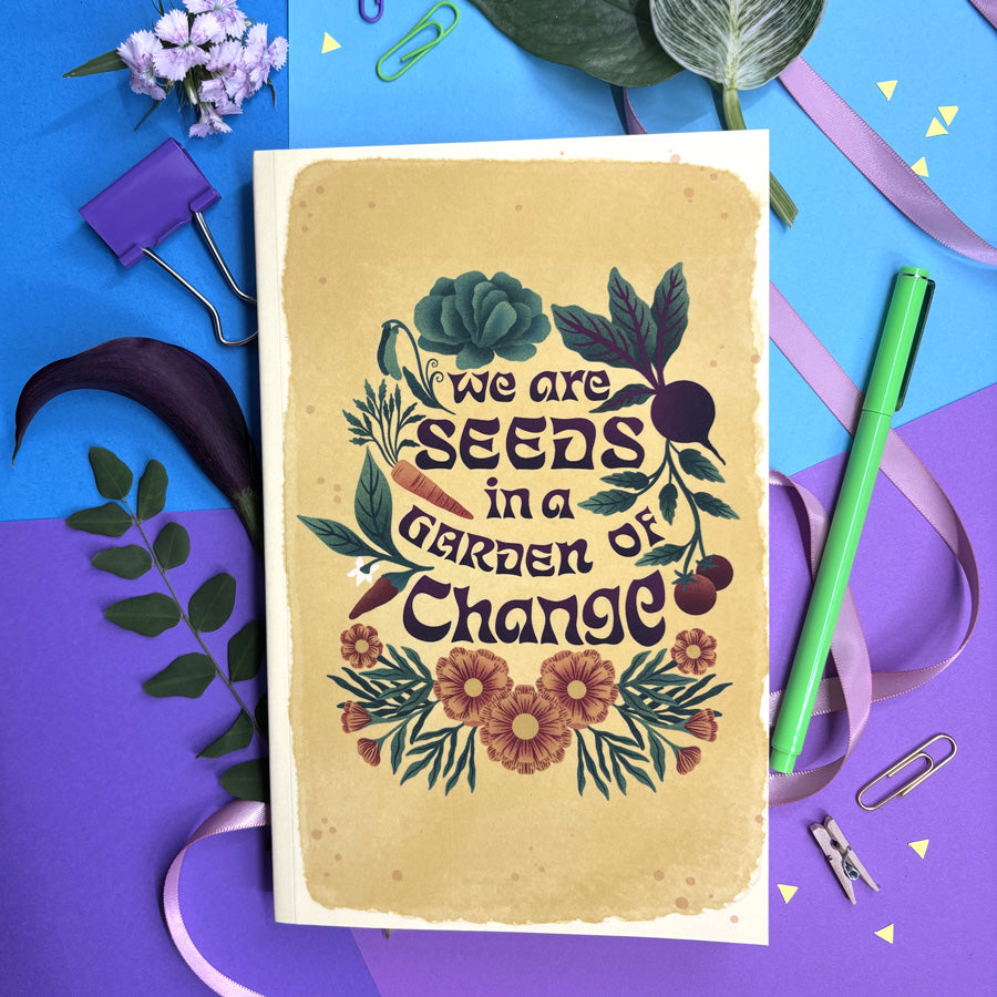 journal with 'We are seeds in a garden of change' text on a colorful background with flowers and stationery items.
