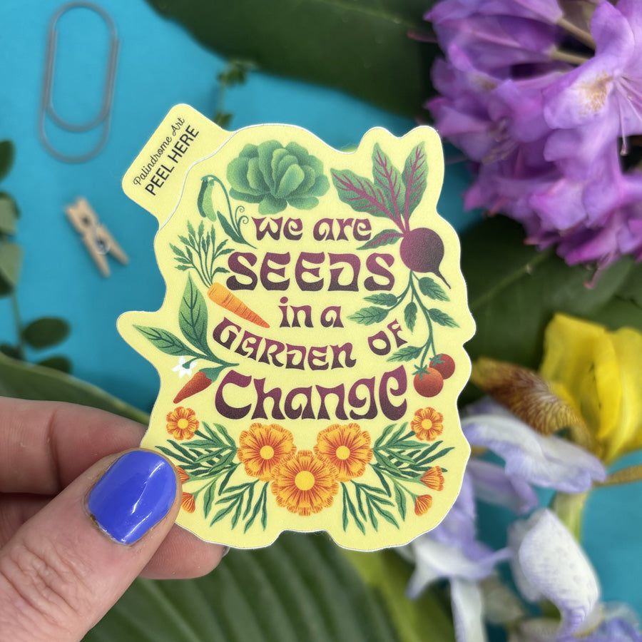 Hand holding a sticker with 'We are seeds in a garden of change' text amidst flowers and greenery.