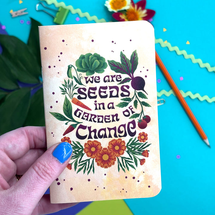 A hand with blue nail polish holdsA small notebook with rounded corners, featuring a warm yellow background and an illustrated design of vegetables, flowers, and the phrase "we are seeds in a garden of change" in dark purple lettering. The background is a bright turquoise surface with a pencil, paper clips, and decorative paper accents.