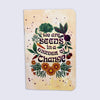 A small notebook with rounded corners, featuring a warm yellow background and an illustrated design of vegetables, flowers, and the phrase "we are seeds in a garden of change" in dark purple lettering.