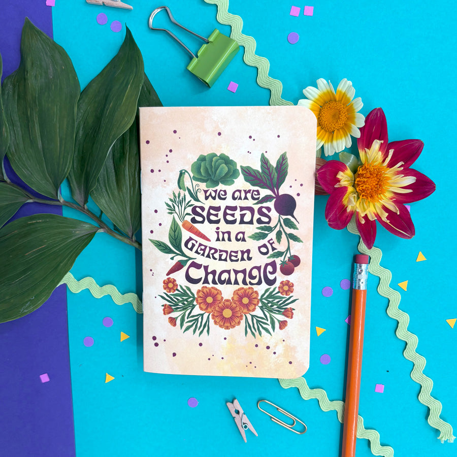 A small notebook with rounded corners, featuring a warm yellow background and an illustrated design of vegetables, flowers, and the phrase "we are seeds in a garden of change" in dark purple lettering is styled in a flat lay on a bright blue background with flowers, a pencil, binder clips, paper clips, and decorative paper accents.