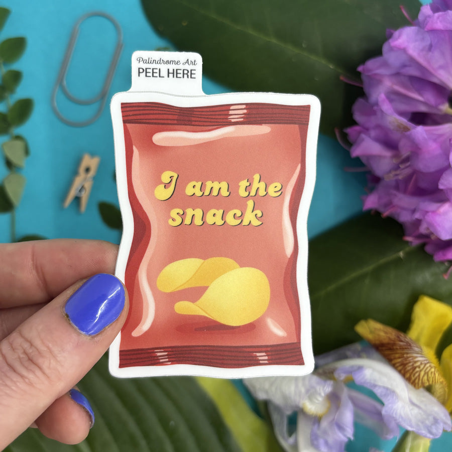Hand holding a sticker with an illustration of a small snack packet with 'I am the snack' text against a colorful background with flowers and leaves.