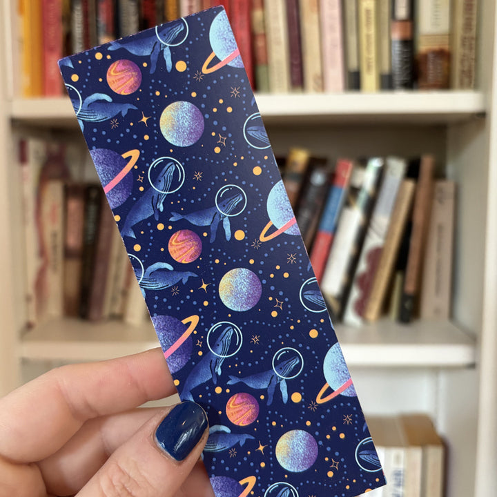 hand holding a bookmark with illustrated whales exploring planets and stars in space in front of a bookshelf