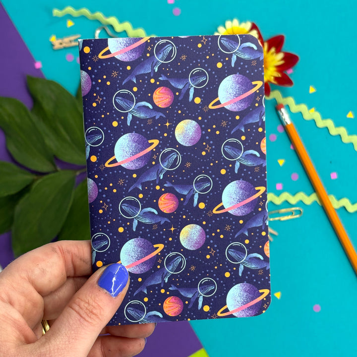 a hand holding a small notebook with rounded corners featuring illustrations of whales with space helmets exploring space amid planets and stars