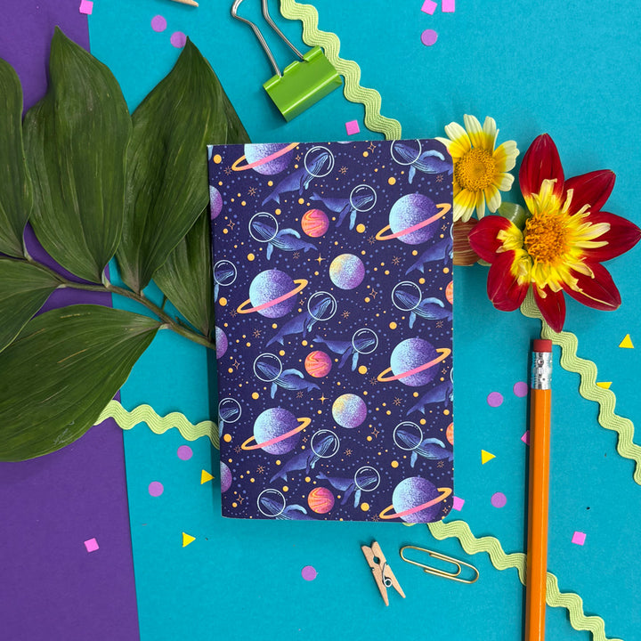 a small notebook with rounded corners featuring illustrations of whales with space helmets exploring space amid planets and stars on a bright background with flowers and stationery items