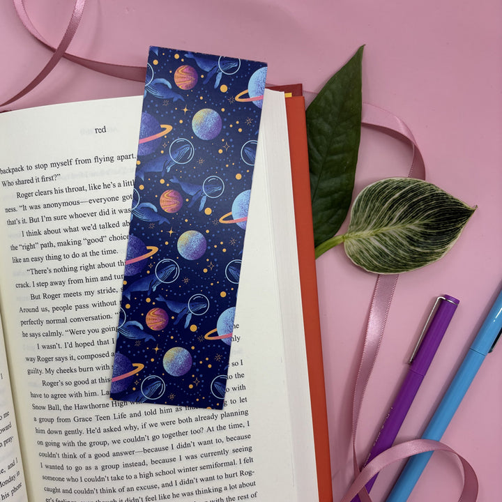 bookmark with illustrated whales exploring planets and stars in space on an open book with a pink background