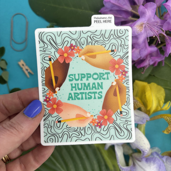sticker with illustration of hands holding pencils and flowers, text 'Support Human Artists', held by a hand against a colorful background.