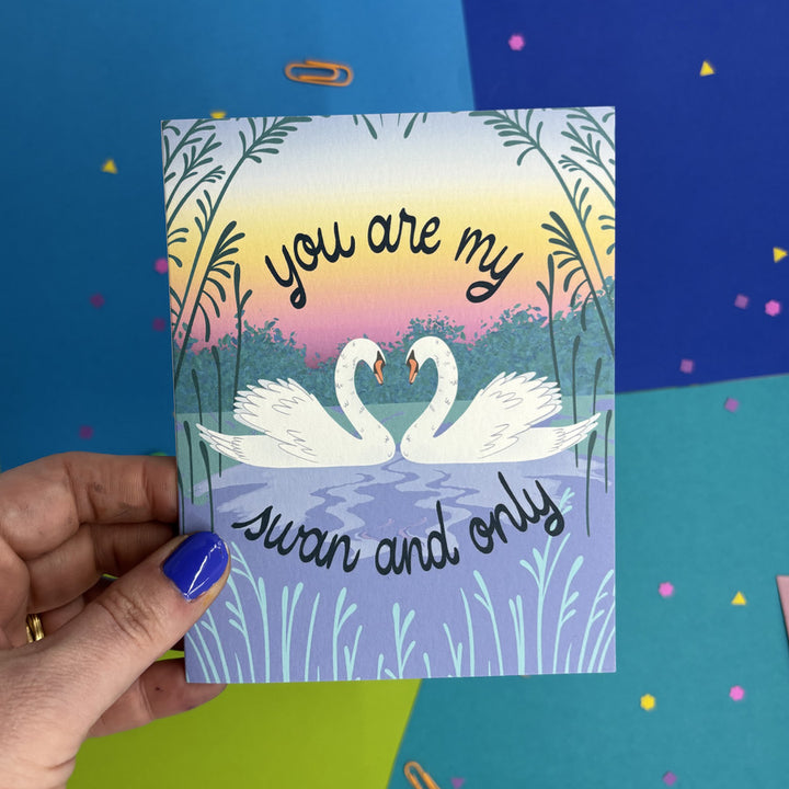 hand holding a greeting card with illustrated swans whose necks make a heart and text "you are my swan and only"