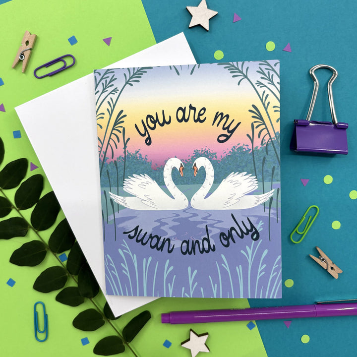 greeting card with illustrated swans whose necks make a heart and text "you are my swan and only" on a colorful background ith stationery items