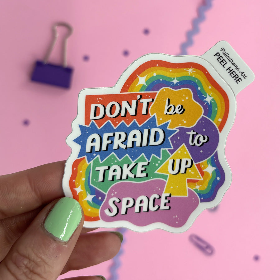 A hand with mint green nail polish holds a die-cut sticker featuring a colorful rainbow and the text 'DON'T be AFRAID to TAKE UP SPACE' in bold white lettering. The sticker has a white peel-and-stick tab in the top right that reads 'PEEL HERE'.