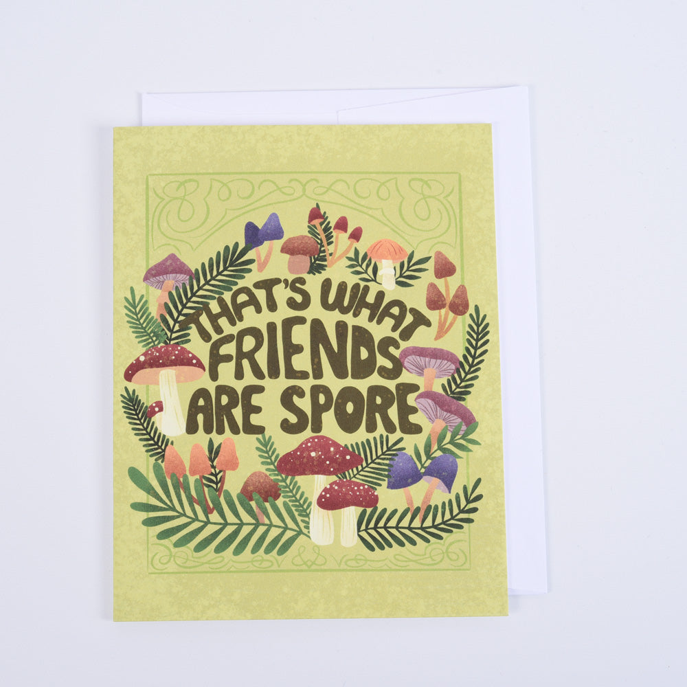 A folded greeting card with a colorful illustration of mushrooms and the text 'That's what friends are spore' on a green background, paired with a bright white envelope.