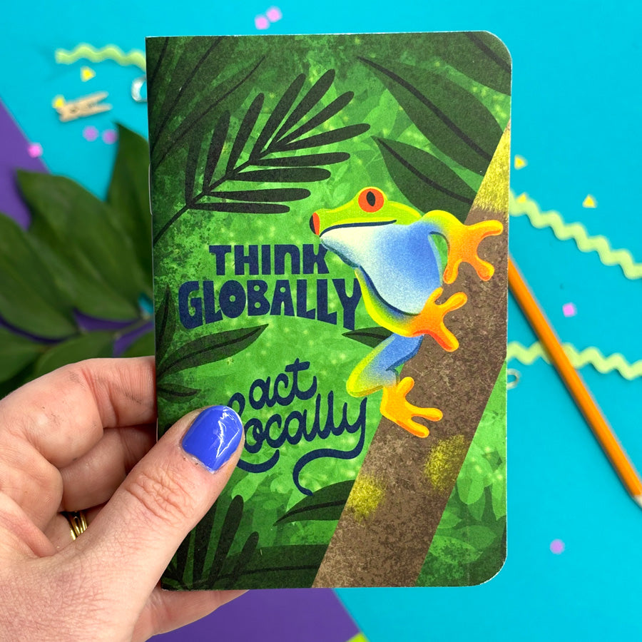 a hand holding a small notebook with rounded corners featuring an illustration of a frog on a thick branch in the jungle with blue lettering "think globally act locally" in front of a bright background