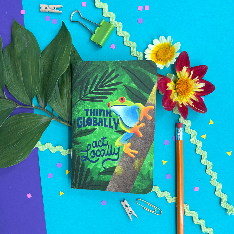 a small notebook with rounded corners featuring an illustration of a frog on a thick branch in the jungle with blue lettering "think globally act locally" on a background with flowers and stationery items