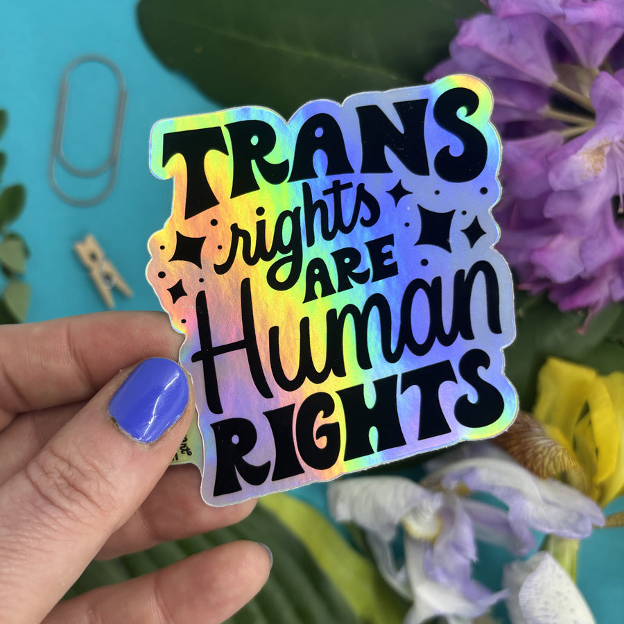Holographic sticker with 'Trans rights are human rights' text held by a hand against a floral background.