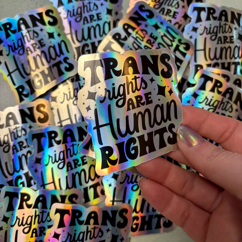 hand holding a holographic sticker that says "trans rights are human rights" with other stickers in the background