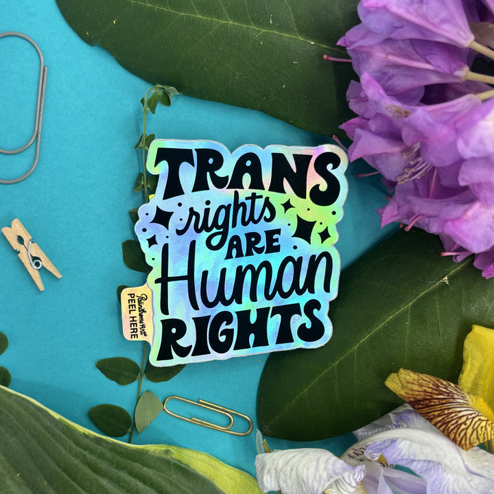 Holographic sticker with 'Trans rights are human rights' text on a blue background with flowers and leaves.