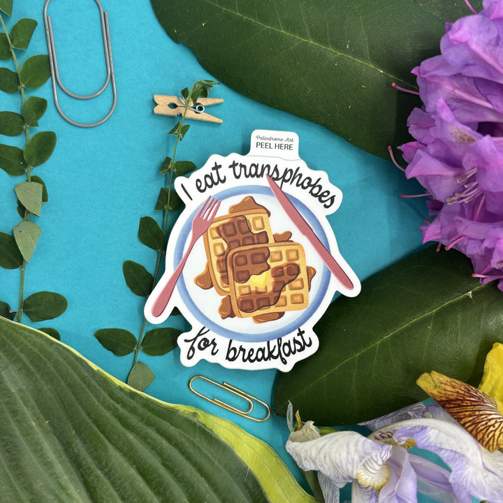Sticker with waffles and text "I eat transphobes for breakfast" on a blue background with leaves and flowers