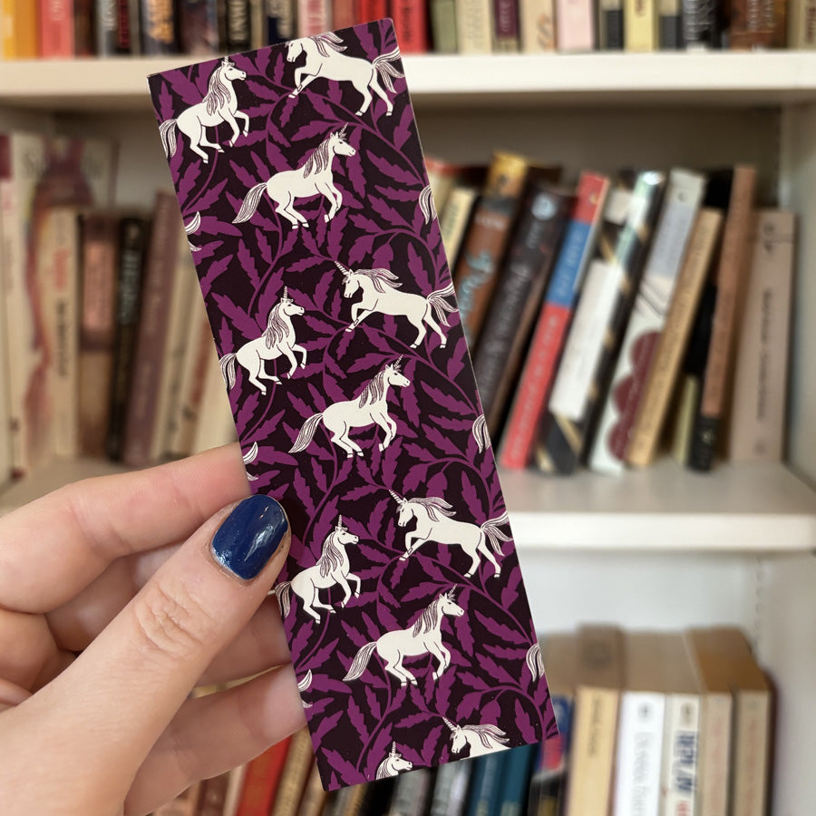 Hand holding a purple bookmark with unicorn pattern in front of a bookshelf.