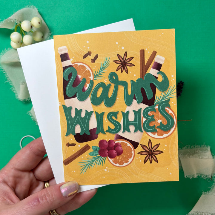 Greeting card with 'Warm Wishes' text and festive illustrations held by a hand on a green background.