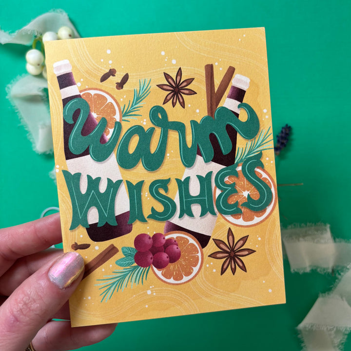 Hand holding a card with 'Warm Wishes' text and festive illustrations on a green background.