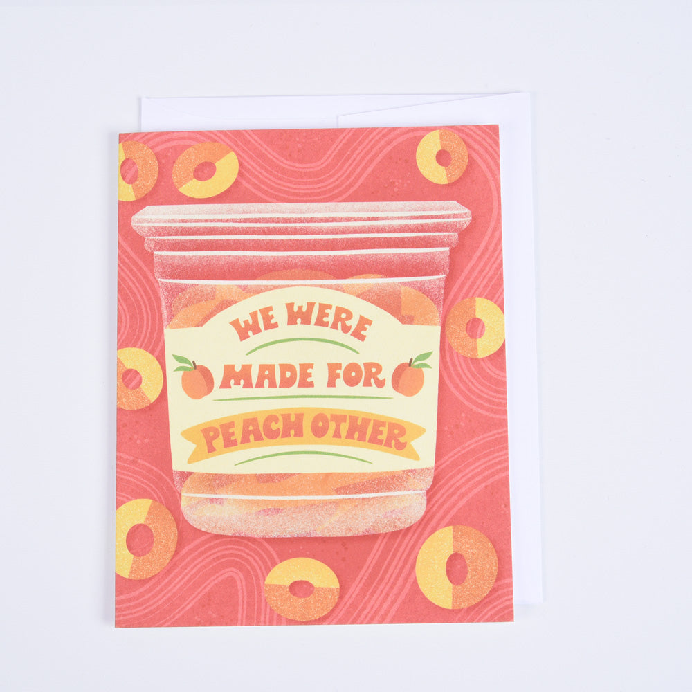 A folded greeting card with an illustration of a plastic container of peach gummy candies and message 'We Were Made for Peach Other' , paired with a bright white envelope.