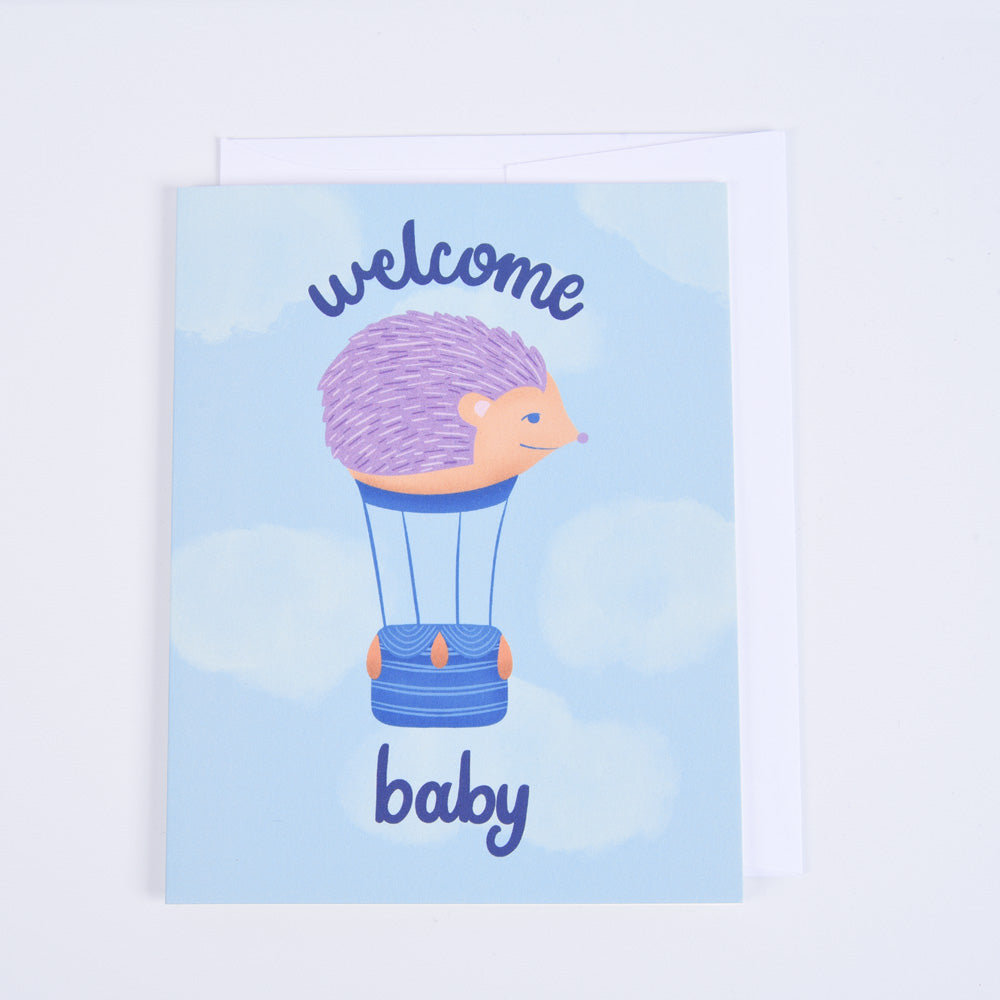 A greeting card with a blue background featuring an illustration of a hedgehog in a hot air balloon, with the text 'Welcome Baby' 
