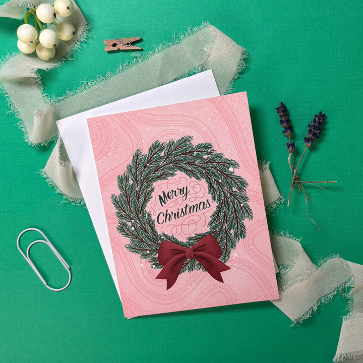 Christmas card with wreath and bow on pink background, surrounded by decorative items on a green surface.