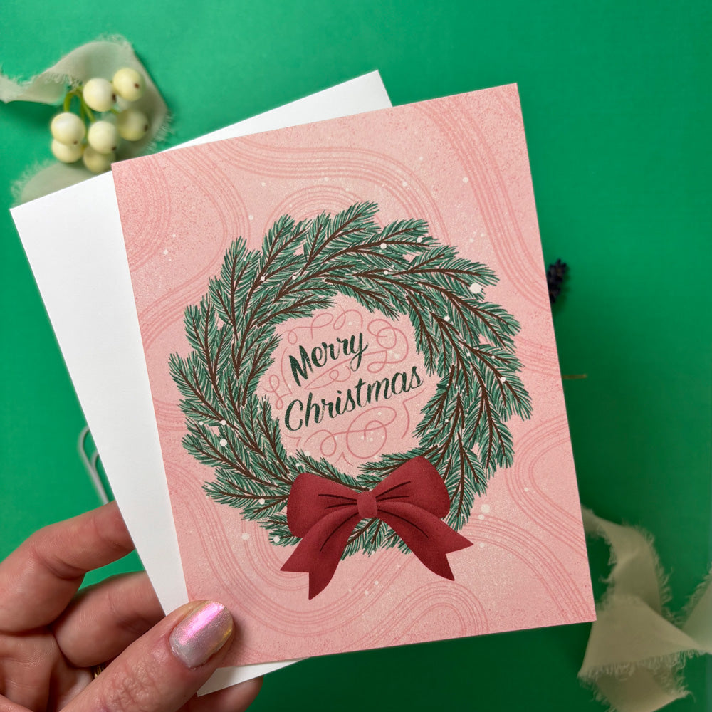 Hand holding a pink Christmas card with a wreath and bow design on a green background.