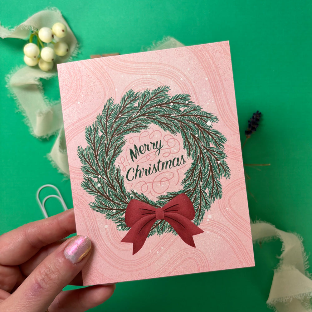 Pink Christmas card with green wreath and red bow held by a hand on a green background