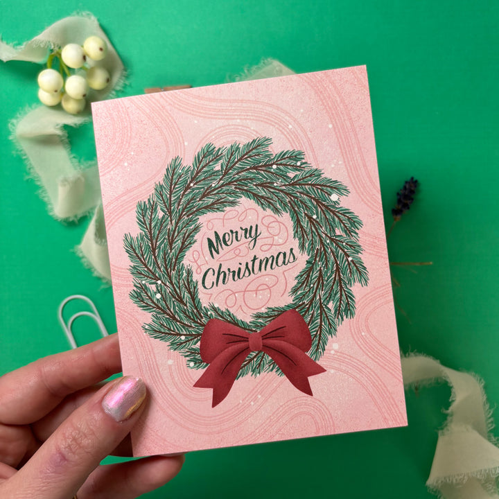 Pink Christmas card with green wreath and red bow held by a hand on a green background
