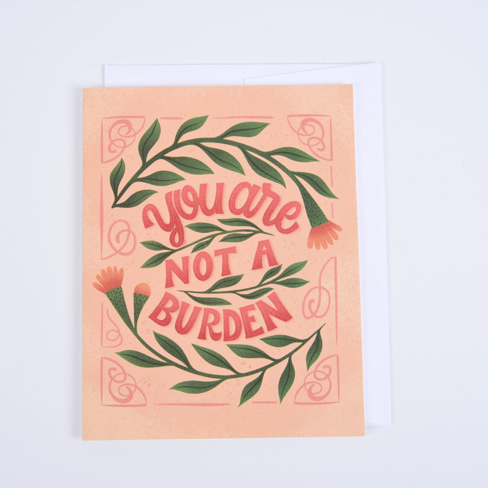 A folded greeting card with a floral pattern and the message 'You Are Not a Burden' printed on the front, accompanied by a bright white envelope.