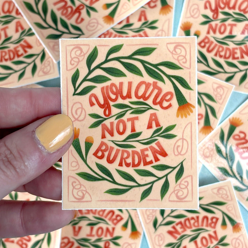 hand holding A sticker with a floral pattern and the text 'You are not a burden' in a red cursive font, on a pink background.