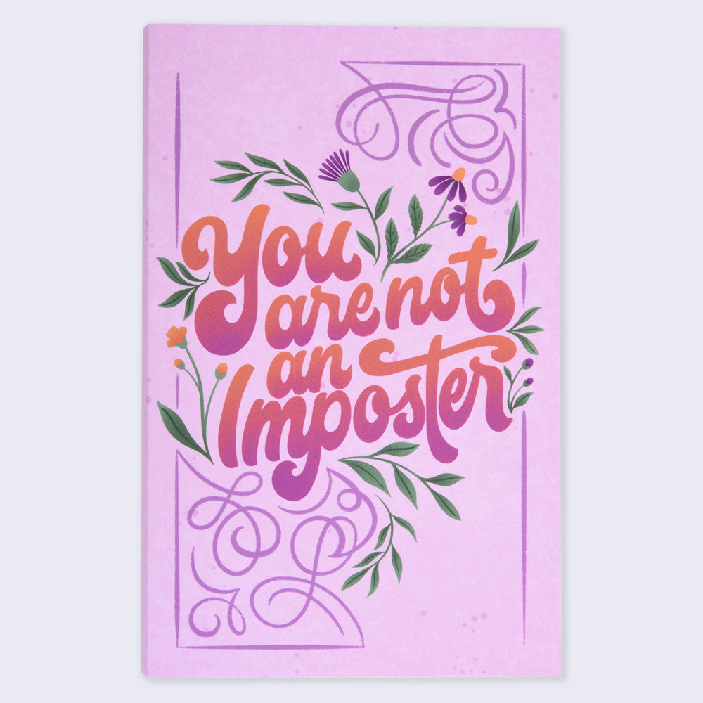 A flat lay view of a pink notebook with a floral design and the text 'You are not an imposter' in hand lettering
