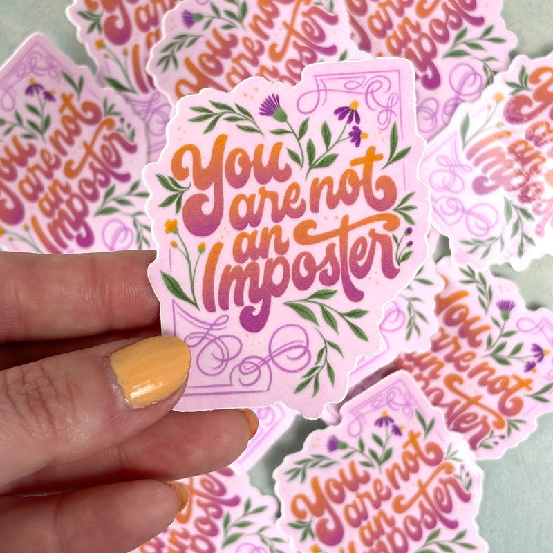 handing holding A sticker with a floral design and the lettering 'you are not an imposter' 